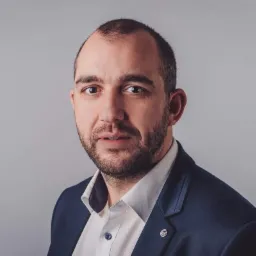 Portret mężczyzny w ciemnym garniturze i jasnej koszuli na szarym tle, z widoczną przypinką z inicjałami na klapie.