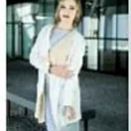 Kobieta w jasnej sukience i białym płaszczu, pozująca na tle nowoczesnego budynku z przeszkleniami. Ujęcie z dołu, podkreślające sylwetkę.