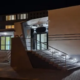 Oświetlona fasada budynku po zmroku z dwoma wejściami, schody z metalowymi barierkami i oświetleniem punktowym, elewacja z poziomymi pasami.