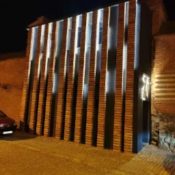 Adaptacja baszty na toaletę miejską w Lęborku: nocne ujęcie elewacji z cegły z pionowymi elementami oświetlonymi od góry, obok zaparkowany czerwony samochód.