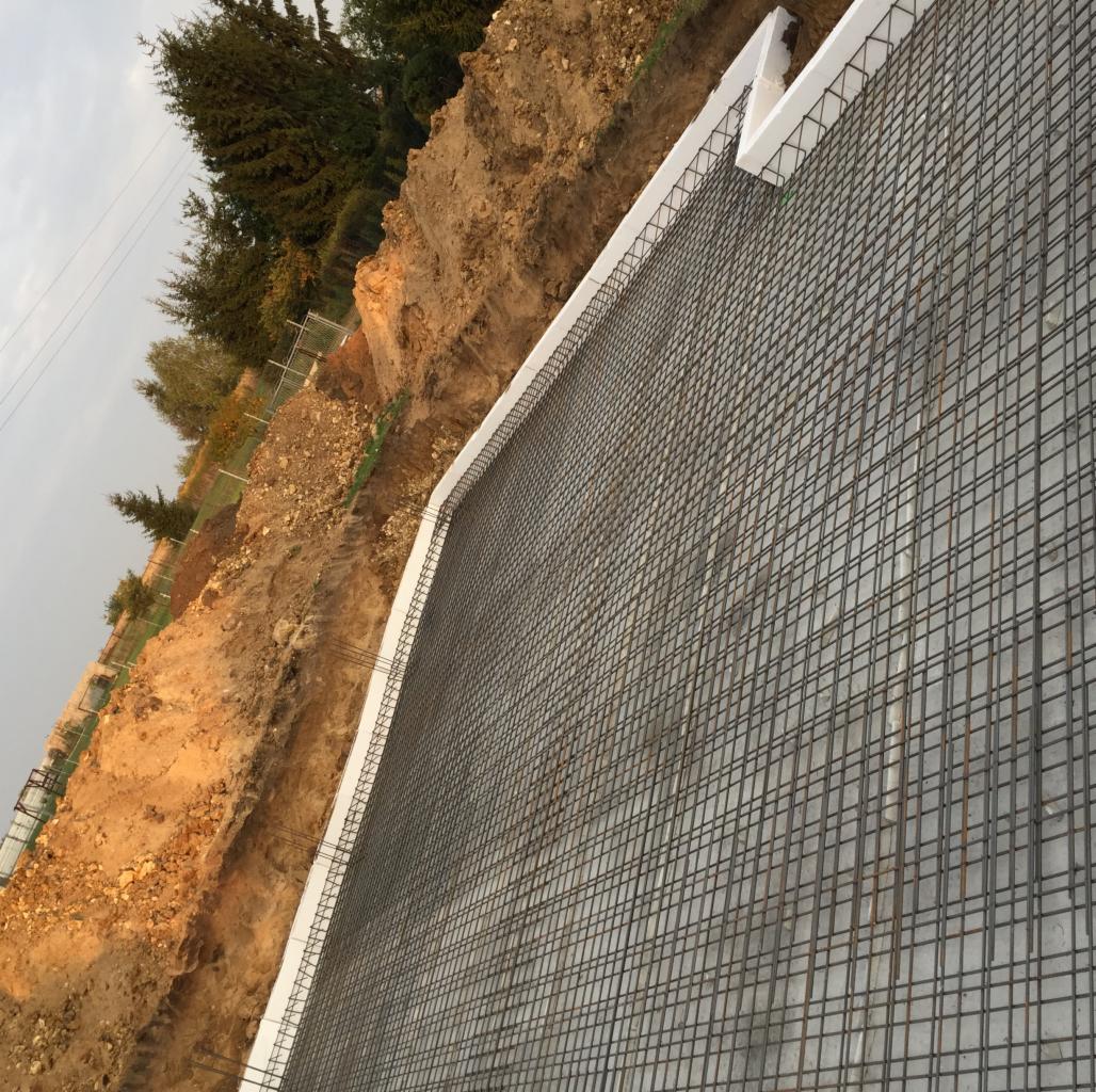 Siatka zbrojeniowa na betonowej ścianie oporowej, zabezpieczona styropianem, widoczna na tle ziemi i zieleni, w pochmurny dzień.