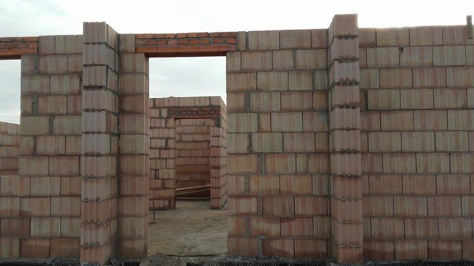 Wznoszenie ścian z bloczków ceramicznych na budowie domu jednorodzinnego. Widoczne otwory okienne i drzwiowe w surowej konstrukcji, ceglany nadproże.