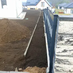 Przygotowany teren pod ogród z hałdą ziemi, widoczny fragment ogrodzenia, obrzeża trawnikowe i domy w tle.