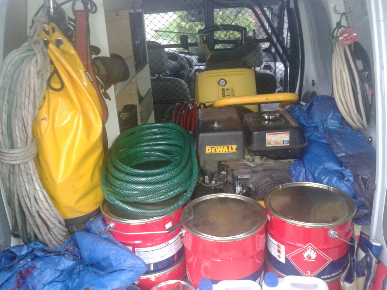 Wnętrze furgonetki z narzędziami: liny, żółty worek, zwój zielonego węża, kanistry i urządzenie DeWalt. Widok z tyłu pojazdu.
