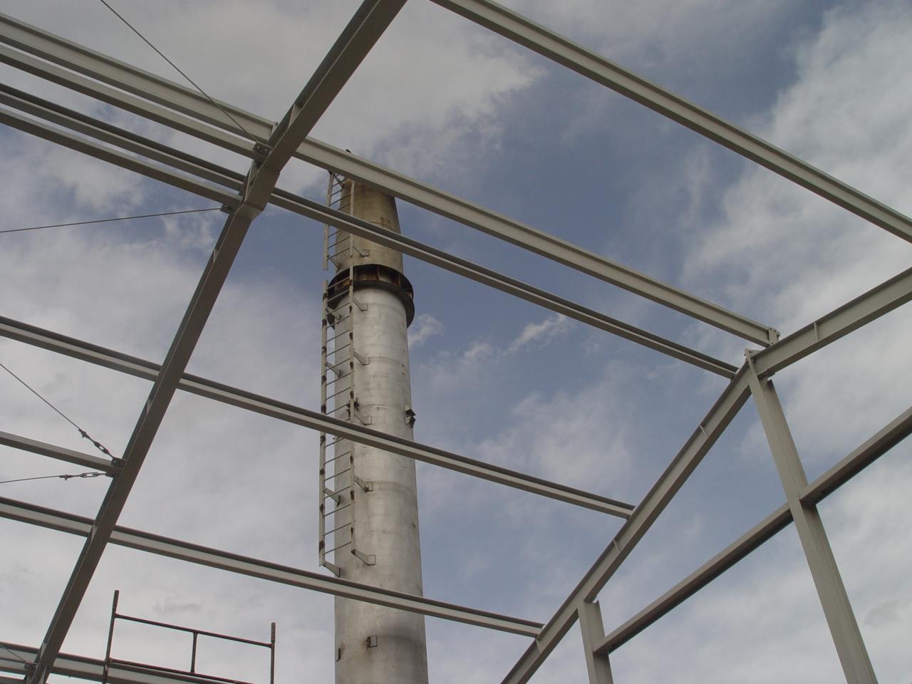 Metalowa konstrukcja dachu w trakcie budowy widziana z dołu, z kominem w tle i fragmentem nieba. Elementy złączone śrubami, widoczne chmury.