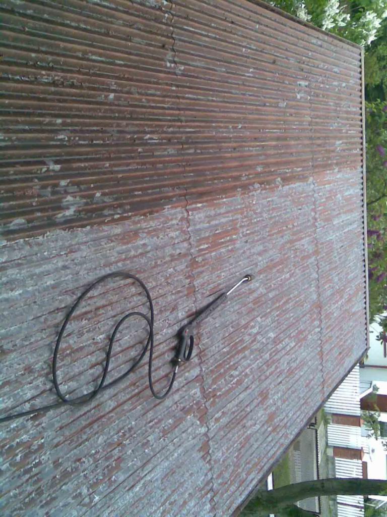 Częściowo wyczyszczony, pordzewiały dach z blachy falistej. Widoczny pistolet do mycia ciśnieniowego i różnica w kolorze między czystą a brudną częścią dachu.