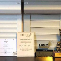 Roletyalu sp. z o.o. - Rolety,Bramy,Kraty,Żaluzje,Okna,Drzwi,Markizy,Pergole,Refleksole. PRODUCENT - Ekspozycja rolet wewnętrznych i zewnętrznych w salonie, z nagrodami i certyfikatami firmy. Widoczne rolety różnych typów i kolorów, dyplomy i statuetki.