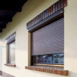 Brązowe rolety zewnętrzne zamontowane na dwóch oknach budynku w Bobrownikach, z dekoracyjną ceglaną obudową nad i pod oknami.