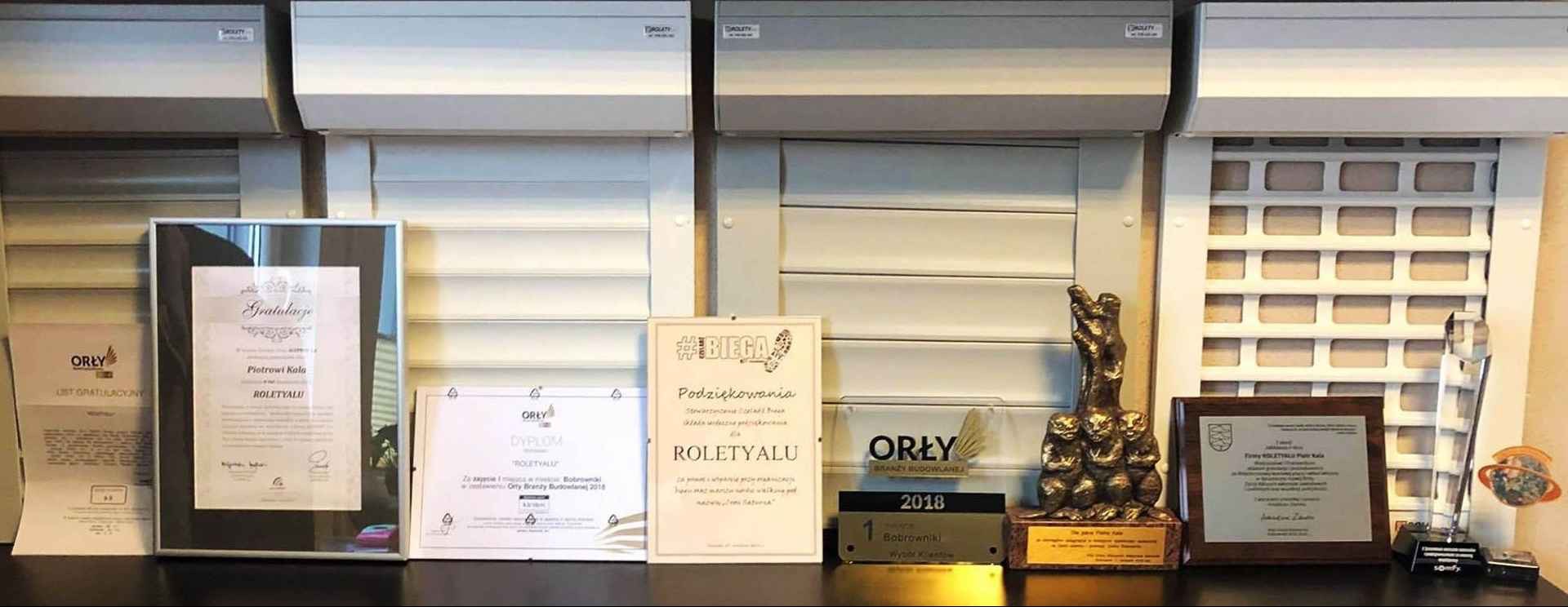 Ekspozycja rolet wewnętrznych i zewnętrznych w salonie, z nagrodami i certyfikatami firmy. Widoczne rolety różnych typów i kolorów, dyplomy i statuetki.