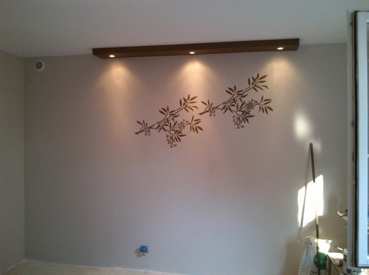 Ściana pomalowana na szaro z naklejonym motywem roślinnym, oświetlona przez reflektory w drewnianej listwie podsufitowej, widoczne akcesoria malarskie i fragment okna.