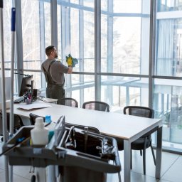 Firma sprzątająca EKOBRIS - Profesjonalne usługi sprzątania - Pracownik w kombinezonie i żółtych rękawicach czyści szmatką duże okno w biurze. W tle widać biurka, krzesła i wózek z akcesoriami do sprzątania.