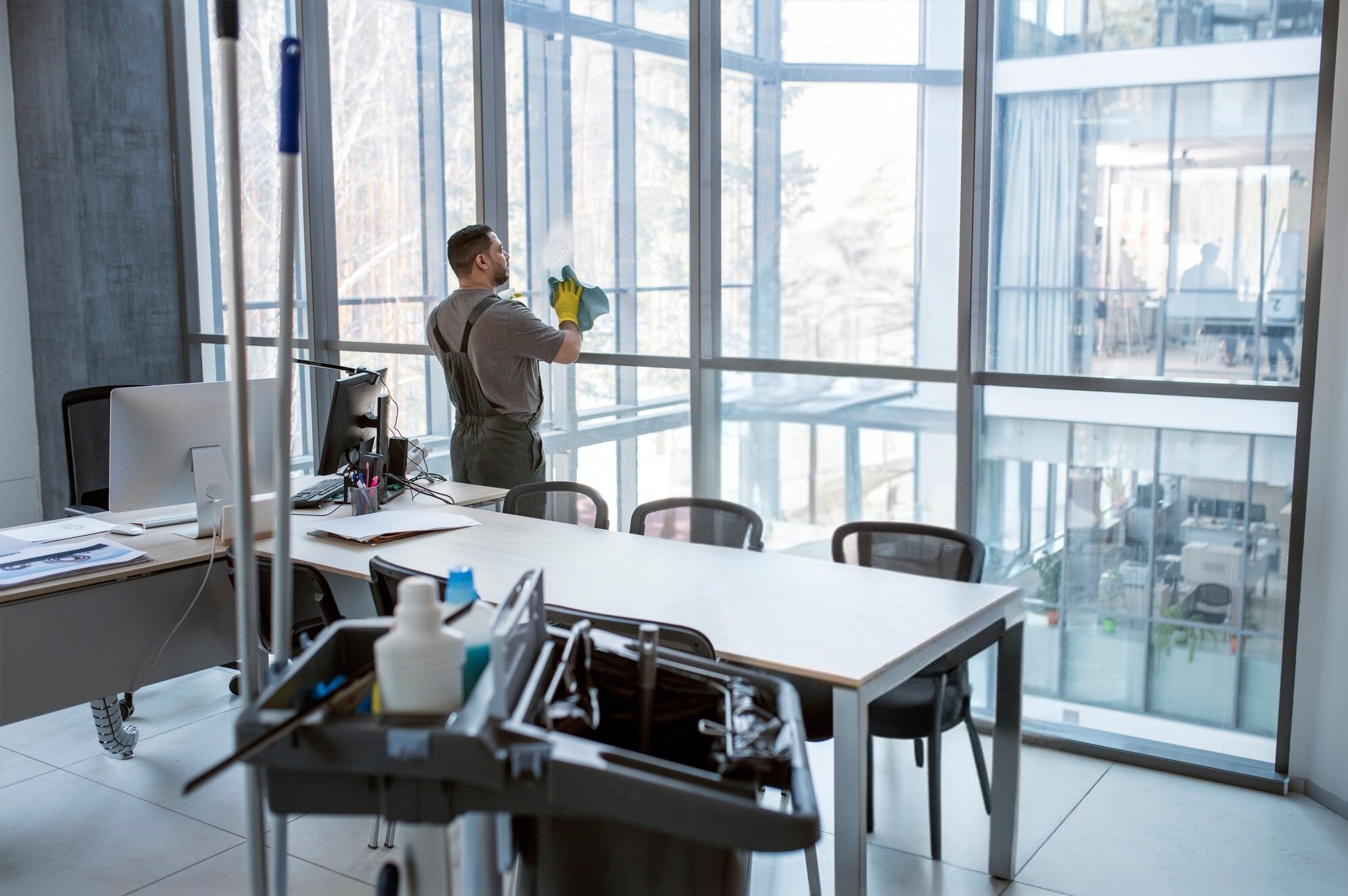Pracownik w kombinezonie i żółtych rękawicach czyści szmatką duże okno w biurze. W tle widać biurka, krzesła i wózek z akcesoriami do sprzątania.