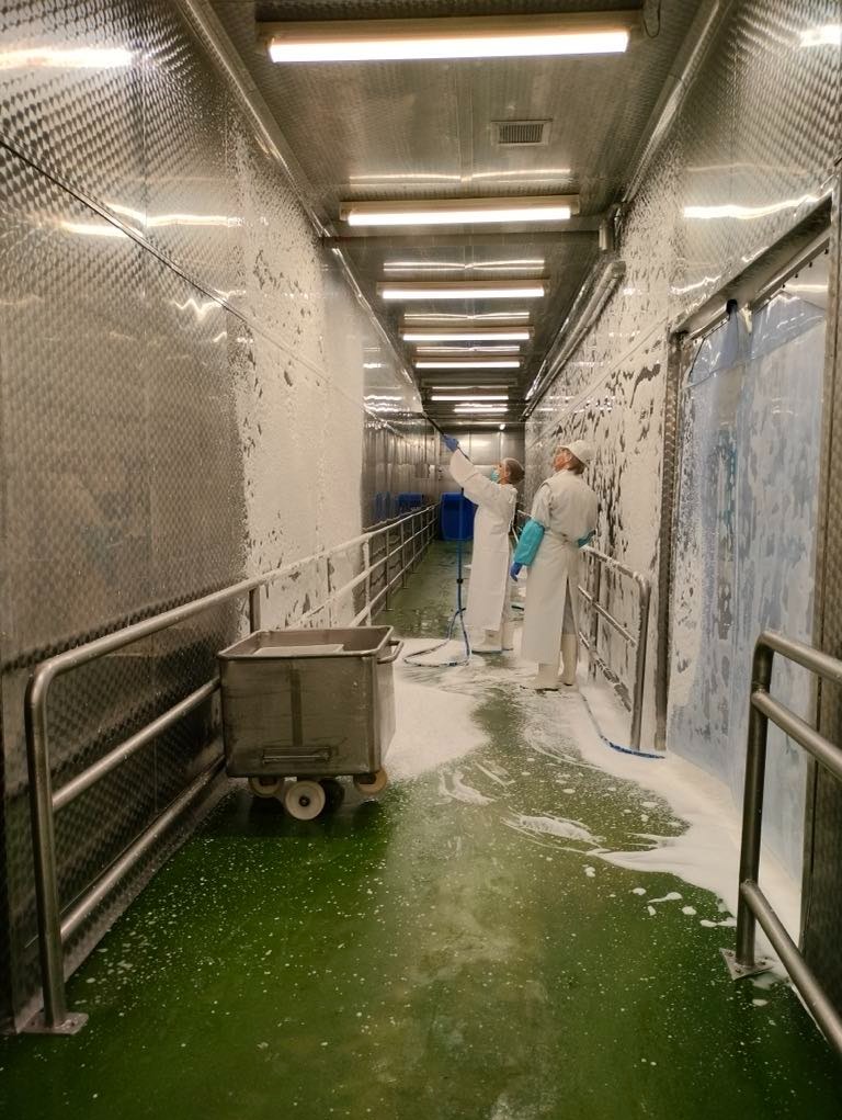 Dwie osoby w kombinezonach ochronnych myją pod ciśnieniem ścianę i podłogę w hali produkcyjnej. Widoczny wózek transportowy i metalowe ściany z fakturą. Na podłodze piana. Oświetlenie jarzeniowe.