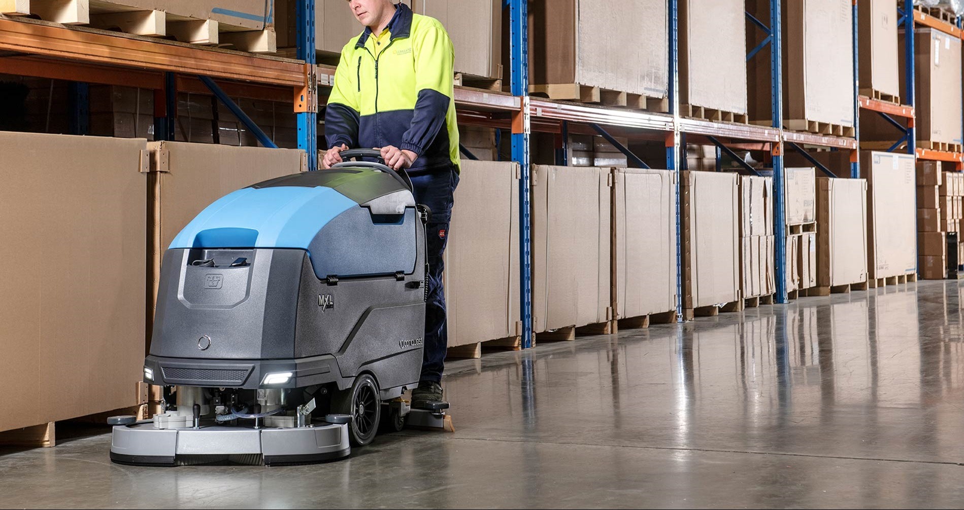 Pracownik w odblaskowej kurtce obsługuje automatyczną szorowarkę do podłóg w magazynie z regałami wypełnionymi kartonami. Maszyna czyszcząca ma niebiesko-szarą obudowę i wbudowane oświetlenie LED.