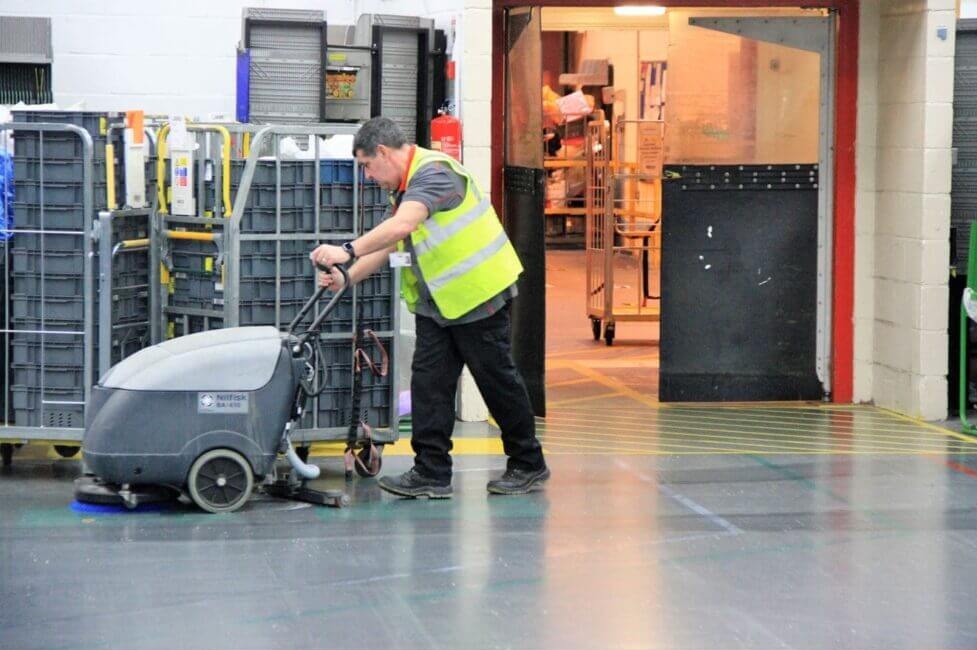 Pracownik w żółtej kamizelce odblaskowej obsługuje szorowarkę Nilfisk BA 450 na błyszczącej, szarej posadzce hali produkcyjnej, w tle regały z pojemnikami i otwarte drzwi.