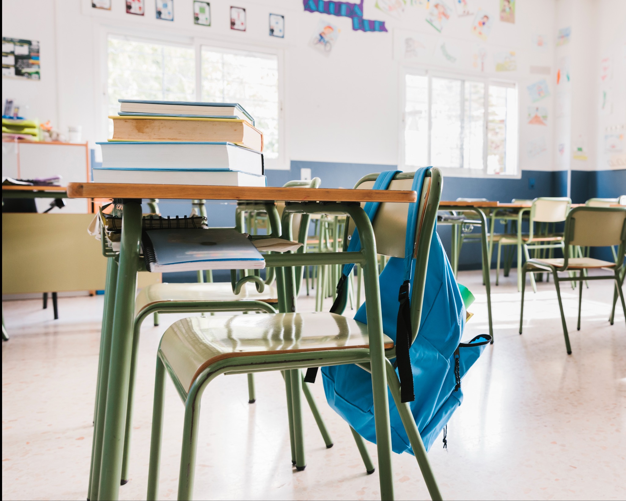 Jasna klasa szkolna z rzędami zielonych ławek i krzeseł, na jednej ławce leżą książki i zeszyty, a na oparciu krzesła wisi niebieski plecak.