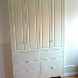 Biała, klasyczna szafa z czterema drzwiami i sześcioma szufladami, z delikatnymi, fioletowymi akcentami na uchwytach, stoi na parkiecie w pokoju.