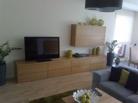 Jasne drewniane meble w salonie: długa komoda pod telewizorem, szafka wisząca, sofa z poduszkami. Uzupełniające dekoracje: rośliny, wazony.