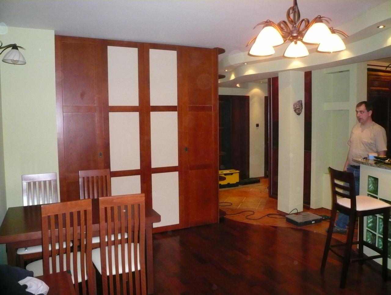 Szafa wnękowa z przesuwnymi drzwiami w odcieniach brązu i beżu, dopasowana do wnętrza z drewnianą podłogą i stołem. Widoczny mężczyzna w tle.