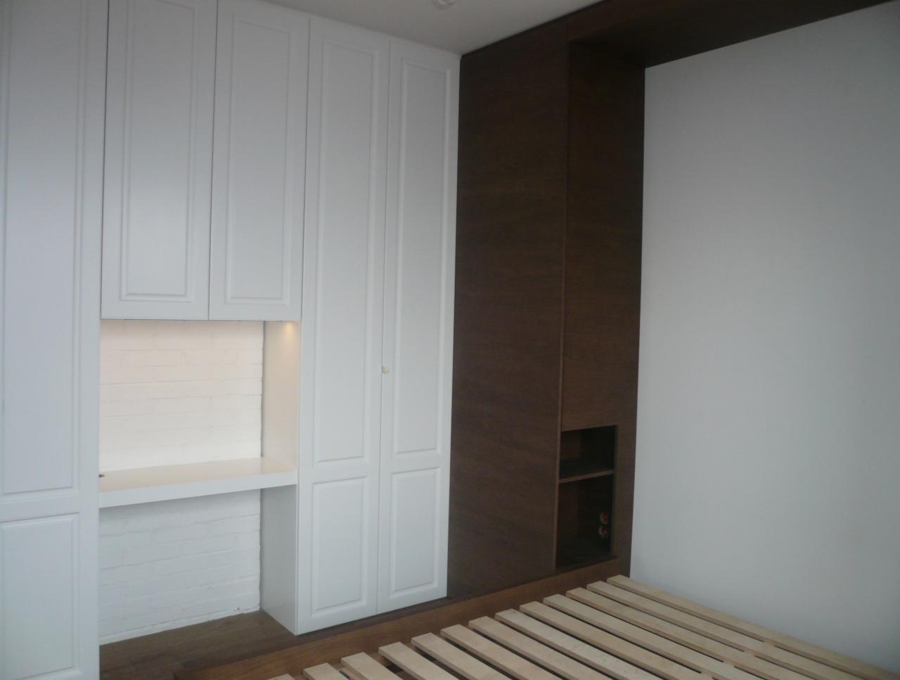 Biała szafa wnękowa z wbudowanym biurkiem i oświetleniem LED, połączona z ciemnobrązową zabudową ściany w sypialni, z widocznym stelażem łóżka.