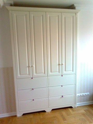 Biała, klasyczna szafa z czterema drzwiami i sześcioma szufladami, z delikatnymi, fioletowymi akcentami na uchwytach, stoi na parkiecie w pokoju.