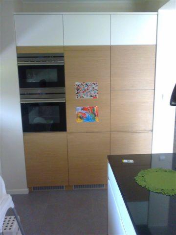 Kuchnia z jasnego drewna z wbudowanym piekarnikiem i białymi szafkami górnymi. Na frontach szafek wiszą dziecięce rysunki.