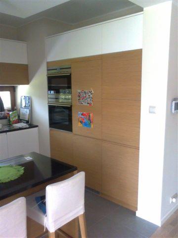 Kuchnia z zabudową z forniru dębowego i białymi szafkami górnymi. Na frontach szafek wiszą dziecięce rysunki. Widoczny stół z czarnym blatem i białe krzesła.