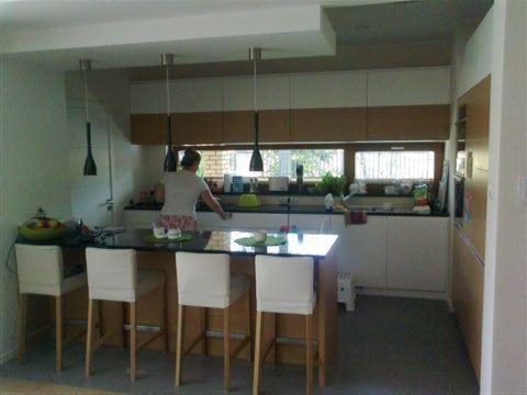 Nowoczesna kuchnia z białymi szafkami i drewnianymi akcentami, wyspą z czarnym blatem i białymi hokerami. Osoba przy zlewie, okno z widokiem na zieleń.