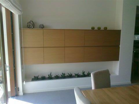 Jasne, drewniane szafki na ścianie z wbudowaną półką na rośliny, nowoczesny design, minimalistyczny styl. Dekoracyjne elementy na górze szafek.