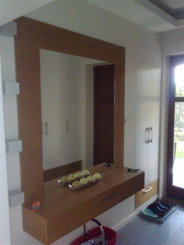 Lustro w drewnianej ramie z blatem i szufladą, zamontowane na ścianie w przedpokoju, obok szafy wnękowej z jasnymi frontami. Na blacie dekoracyjne owoce.