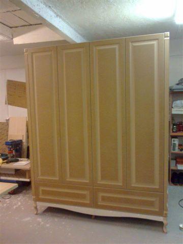 Beżowa, czterodrzwiowa szafa z frezowanymi frontami i dekoracyjnymi nóżkami w warsztacie. Widoczne narzędzia i materiały, co sugeruje etap produkcji mebla na wymiar.