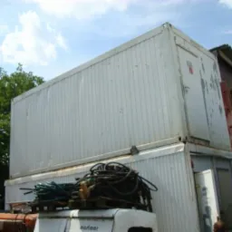 Dwa białe kontenery ustawione jeden na drugim, widoczne przewody i fragmenty pojazdów. Kontenery z widocznymi śladami użytkowania na tle drzew i błękitnego nieba.