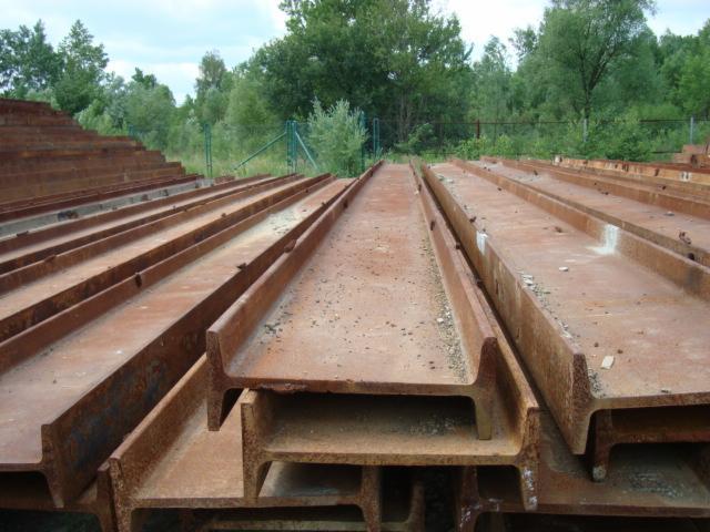 Składowane, zardzewiałe, stalowe belki dwuteowe w plenerze, ułożone równolegle, tworzące schodkową strukturę. W tle zieleń drzew i krzewów.