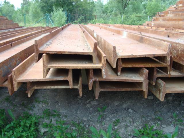 Stalowe, zardzewiałe belki dwuteowe ułożone w stos, widoczne na tle zieleni. Belki o charakterystycznym przekroju, z widoczną fakturą rdzy.