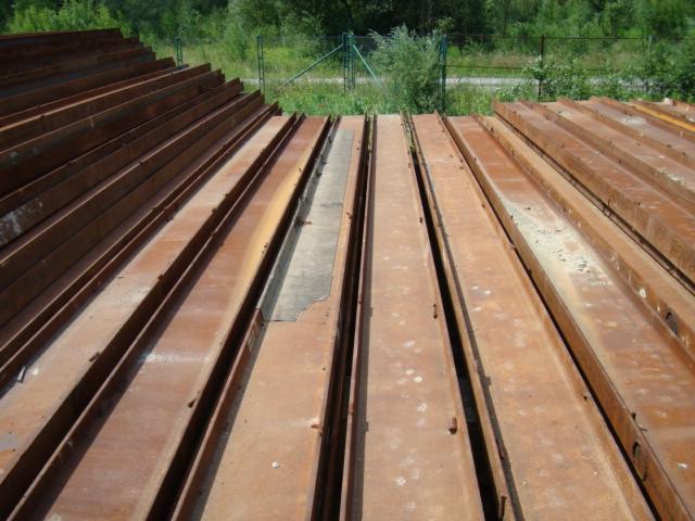 Stalowe, zardzewiałe belki ułożone równolegle, widoczne ślady korozji i zabrudzeń. Tło z zielenią i siatką ogrodzeniową.