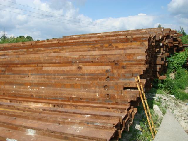 Stos zardzewiałych, metalowych belek o prostokątnym przekroju, ułożonych warstwami na zewnątrz, z zielenią w tle i fragmentem drabiny.