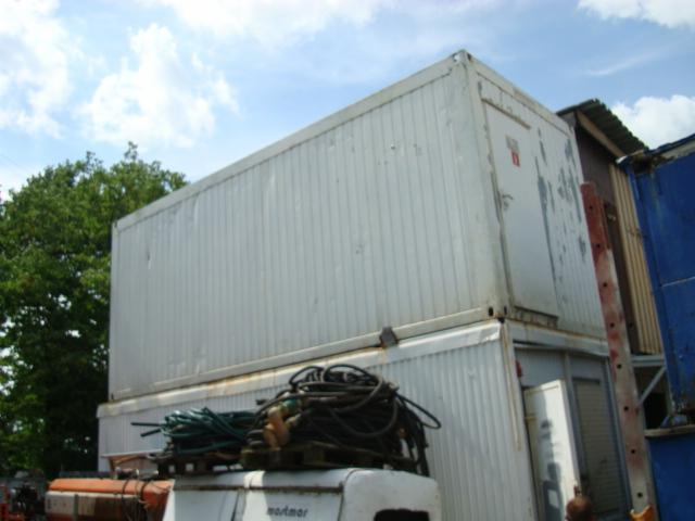 Dwa białe kontenery ustawione jeden na drugim, widoczne przewody i fragmenty pojazdów. Kontenery z widocznymi śladami użytkowania na tle drzew i błękitnego nieba.