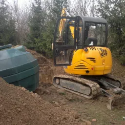 Żółta mini koparka JCB obok zielonego zbiornika na tle ziemi i drzew. Wykopany rów sugeruje instalację przydomowej oczyszczalni ścieków.