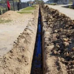 Wykop w ziemi z ułożonymi niebieskimi rurami, widok z góry wzdłuż wykopu, w tle zabudowa jednorodzinna.