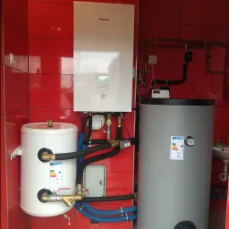 Zestaw instalacji grzewczej z pompą ciepła Panasonic Aquarea, dwoma zbiornikami (białym i szarym), rurami w izolacji i armaturą na tle ściany wyłożonej czerwoną glazurą.