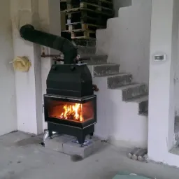 Czarny piec wolnostojący z paleniskiem, podłączony do komina, w surowym wnętrzu z betonowymi schodami i paletami w tle.