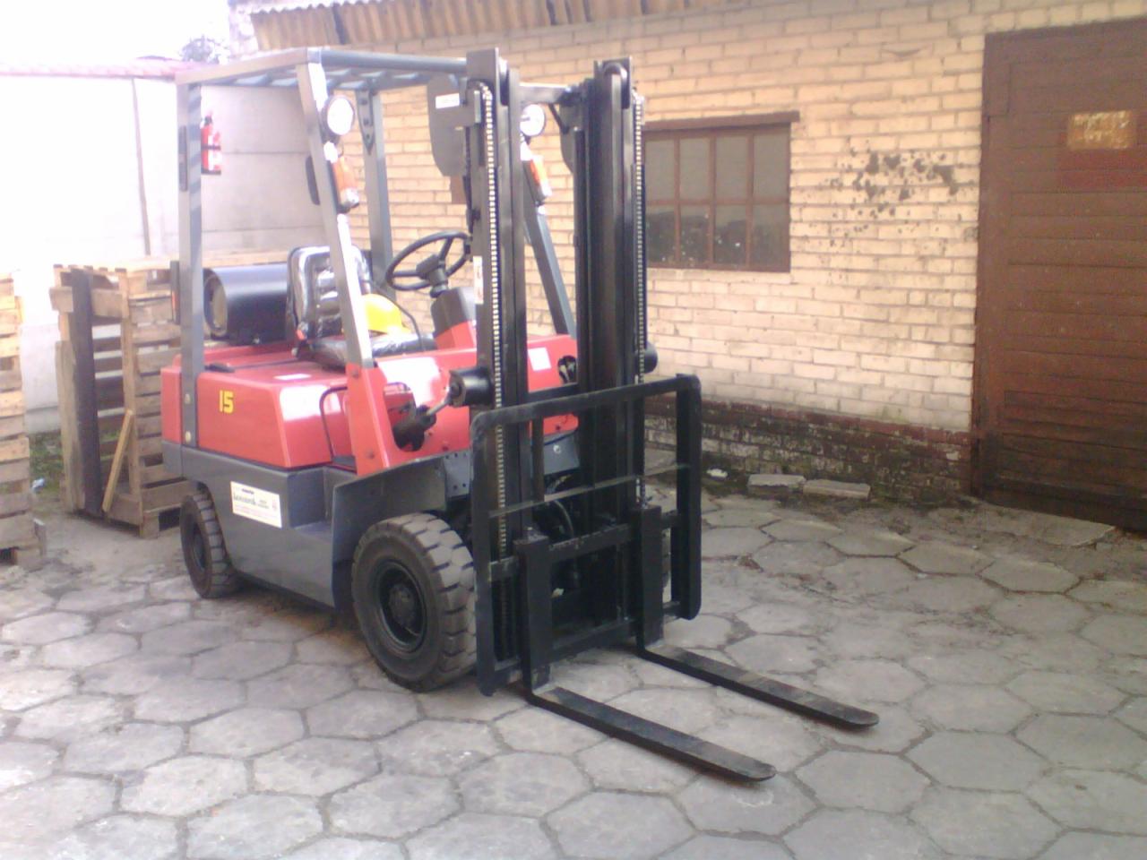 Czerwono-szary wózek widłowy z czarnym osprzętem na zewnątrz, zaparkowany na sześciokątnej kostce brukowej, z paletami w tle i ceglanym budynkiem z brązowymi drzwiami.