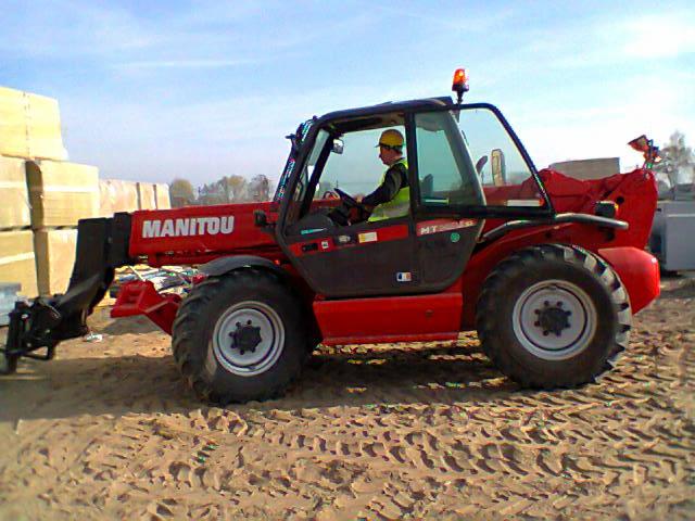 Czerwony wózek widłowy Manitou z operatorem w żółtym kasku, stojący na piaszczystym terenie z widocznymi śladami opon, w tle ułożone materiały budowlane.