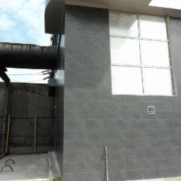 Elewacja budynku z szarą, ceramiczną okładziną, widoczne okno z żaluzjami i fragment instalacji przemysłowej w tle. Całość w dziennym oświetleniu.