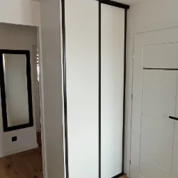 Biała szafa przesuwna z czarnymi obramowaniami w jasnym wnętrzu, widoczna podłoga z paneli i drzwi wewnętrzne.