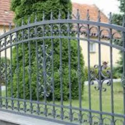 Eleganckie, metalowe ogrodzenie z dekoracyjnymi elementami, pomalowane na szaro, oddziela posesję od trawnika. W tle widoczny budynek i zieleń.