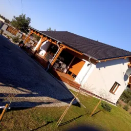 Nowoczesny dom jednorodzinny z ciemną dachówką, drewnianą pergolą i anteną satelitarną, widziany z poziomu trawnika w słoneczny dzień.