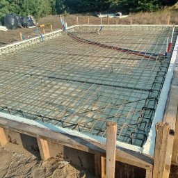 DARBUD Dariusz Zaręba - Zbrojenie płyty fundamentowej w szalunku z izolacją termiczną, przygotowane do zalania betonem, widoczne rury instalacyjne i worki z odpadami w tle.