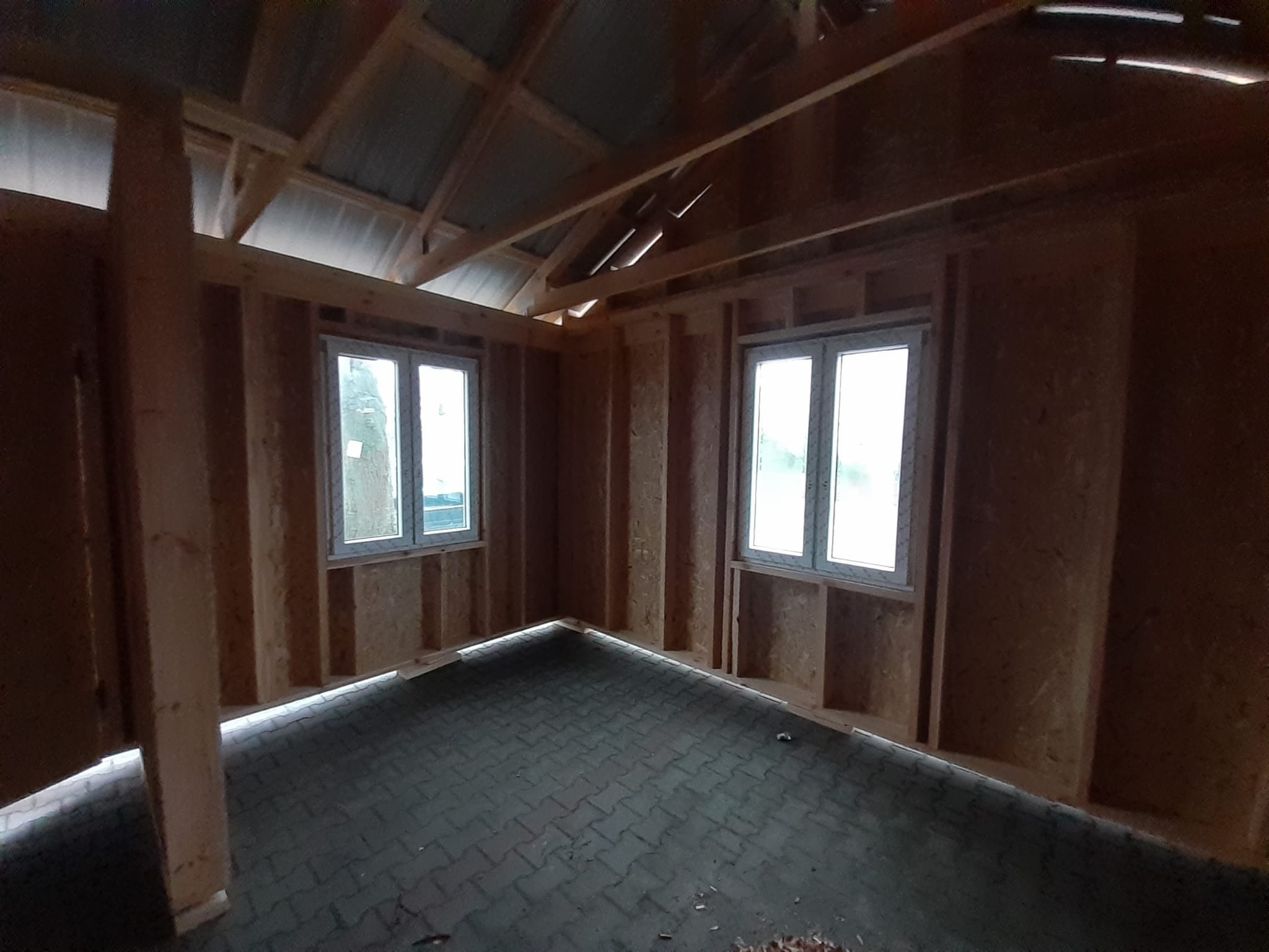 Wnętrze w trakcie budowy: ściany z płyt OSB, drewniana konstrukcja dachu, dwa okna z białymi ramami i podłoga wyłożona szarą kostką brukową.