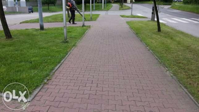 Pracownik z dmuchawą do liści na chodniku z kostki brukowej, trawnik po bokach, ulica w tle. Prace porządkowe na zewnątrz.